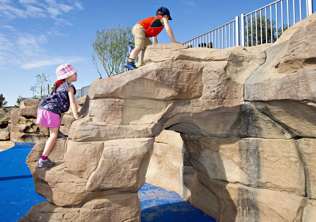 Children play on a sculpted rock formation with an arch, in a bright playground