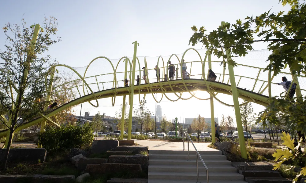 People walk across a curved green playground bridge in a park, framed by trees and warm sunlight.