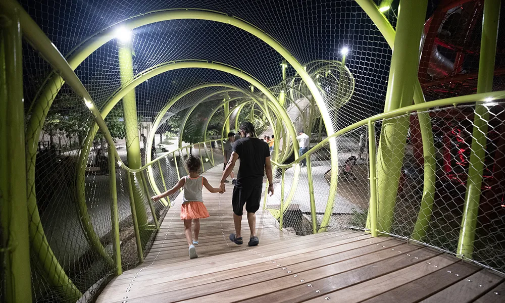 A man and child walk hand in hand through an illuminated green tunnel bridge with circular frames at night.