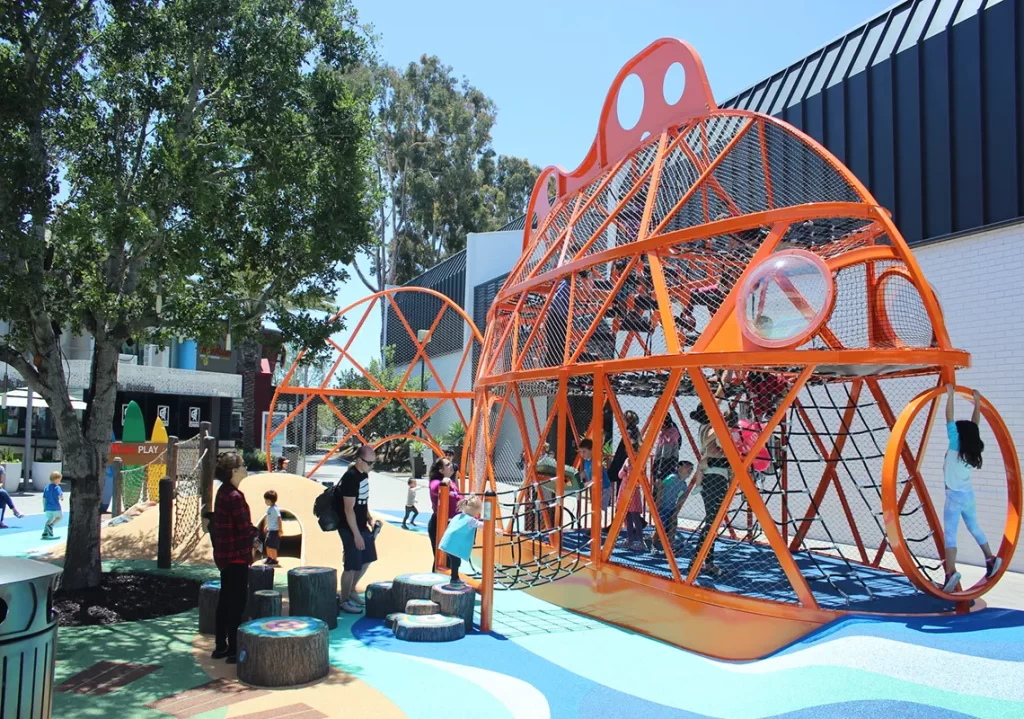 Children play inside a bright orange fish-shaped climbing structure with nets at a colorful outdoor playground