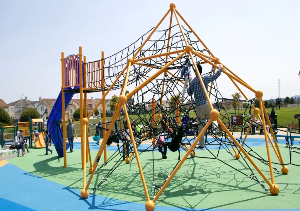 Children climb a large rope frame net with yellow posts in a busy neighborhood playground
